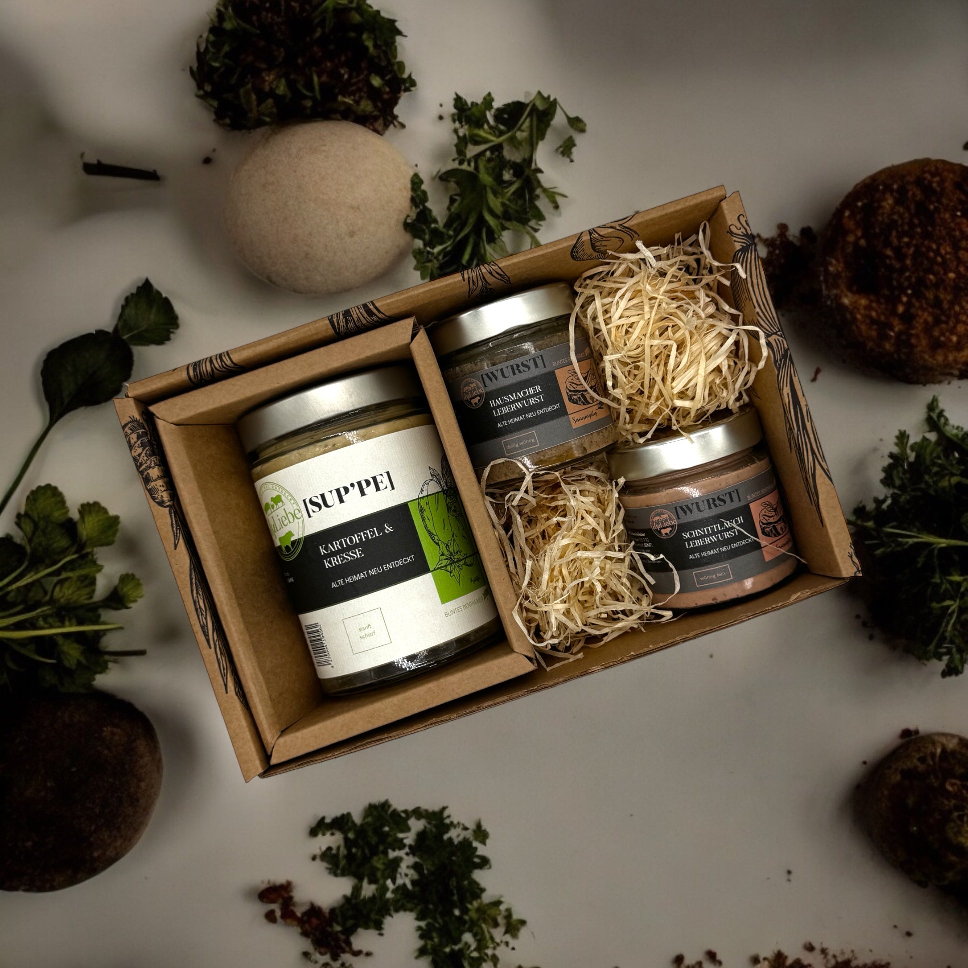 Eine kleine Genussbox der Marke elseLiebe steht auf einer Arbeitsplatte, die dekoriert ist mit Kräutern. In der kulinarischen Präsentbox ist links ein Glas Suppe zu sehen. Rechts liegen auf etwas Holzwolle zwei kleinere Gläser mit Leberwurst vom Bunten Bentheimer Schwein. Es sind die Sorten Hausmacher und Schnittlauch.