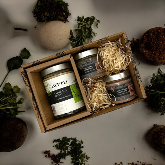 Eine kleine Genussbox der Marke elseLiebe steht auf einer Arbeitsplatte, die dekoriert ist mit Kräutern. In der kulinarischen Präsentbox ist links ein Glas Suppe zu sehen. Rechts liegen auf etwas Holzwolle zwei kleinere Gläser mit Leberwurst vom Bunten Bentheimer Schwein. Es sind die Sorten Hausmacher und Schnittlauch.