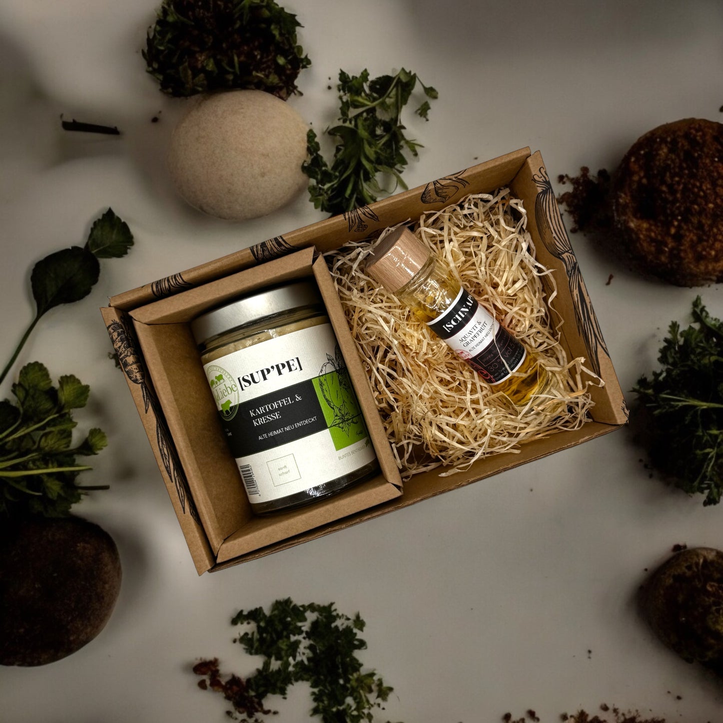 Eine kleine Genussbox der Marke elseLiebe steht auf einer Arbeitsplatte, die dekoriert ist mit Kräutern. In der kulinarischen Präsentbox ist links ein Glas Suppe zu sehen. Rechts liegt auf etwas Holzwolle eine kleine Flasche Schnaps eines Aquavit Likörs mit Grapefruit.