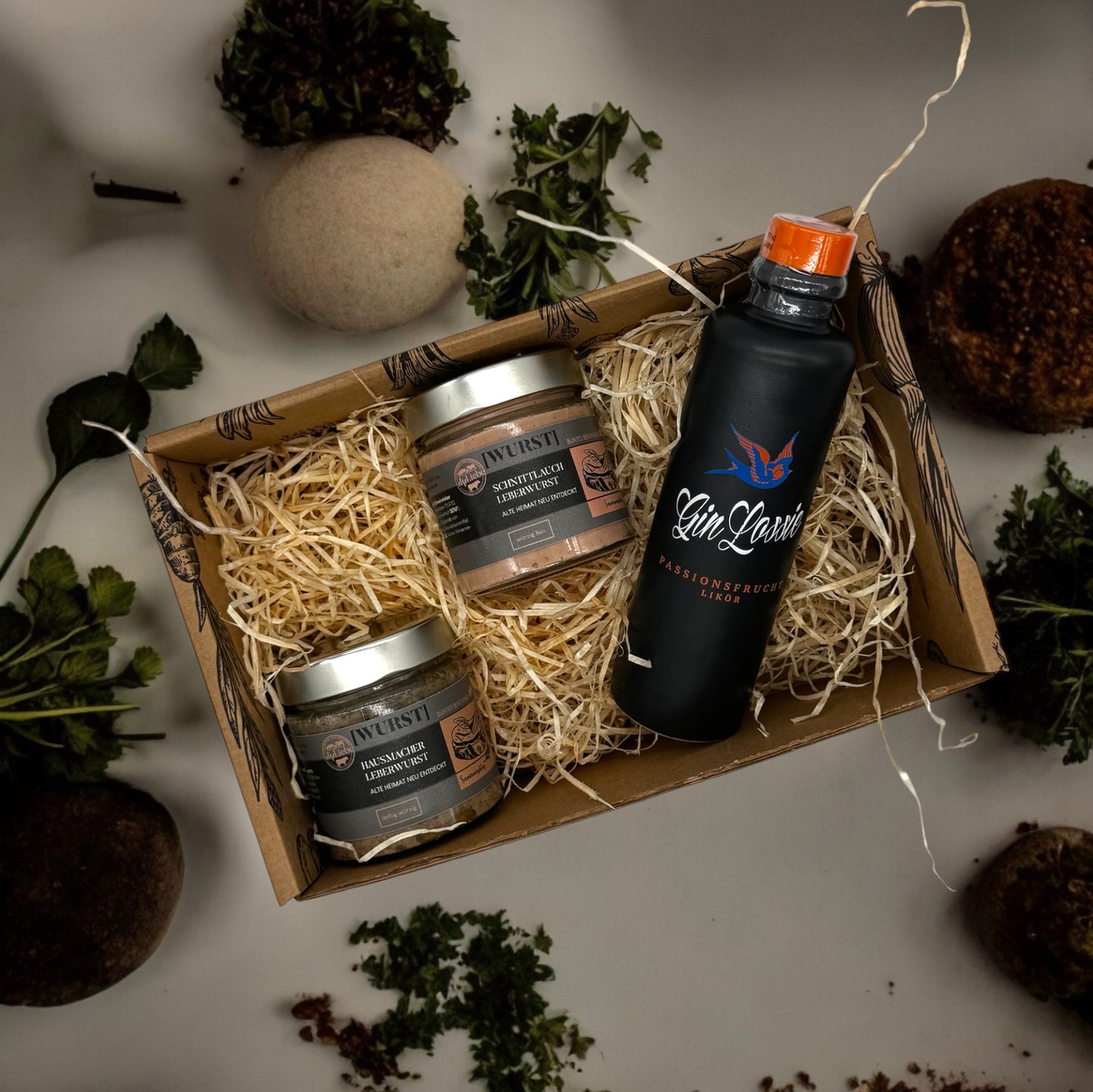 Eine kleine Genussbox der Marke elseLiebe steht auf einer Arbeitsplatte, die dekoriert ist mit Kräutern. Links liegen auf etwas Holzwolle zwei kleine Gläser mit Leberwurst vom Bunten Bentheimer Schwein. Es sind die Sorten Hausmacher und Schnittlauch. Daneben ist eine kleine Flasche Gin Lossie der Sorte Gin Likör Passionsfrucht zu sehen.