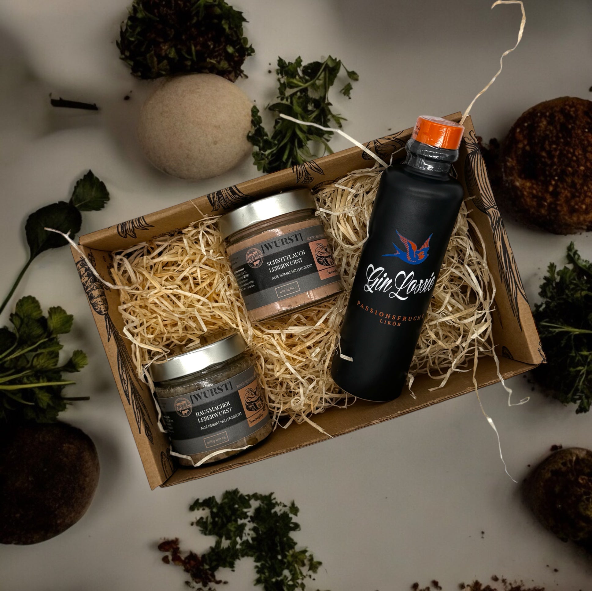 Eine kleine Genussbox der Marke elseLiebe steht auf einer Arbeitsplatte, die dekoriert ist mit Kräutern. Links liegen auf etwas Holzwolle zwei kleine Gläser mit Leberwurst vom Bunten Bentheimer Schwein. Es sind die Sorten Hausmacher und Schnittlauch. Daneben ist eine kleine Flasche Gin Lossie der Sorte Gin Likör Passionsfrucht zu sehen.