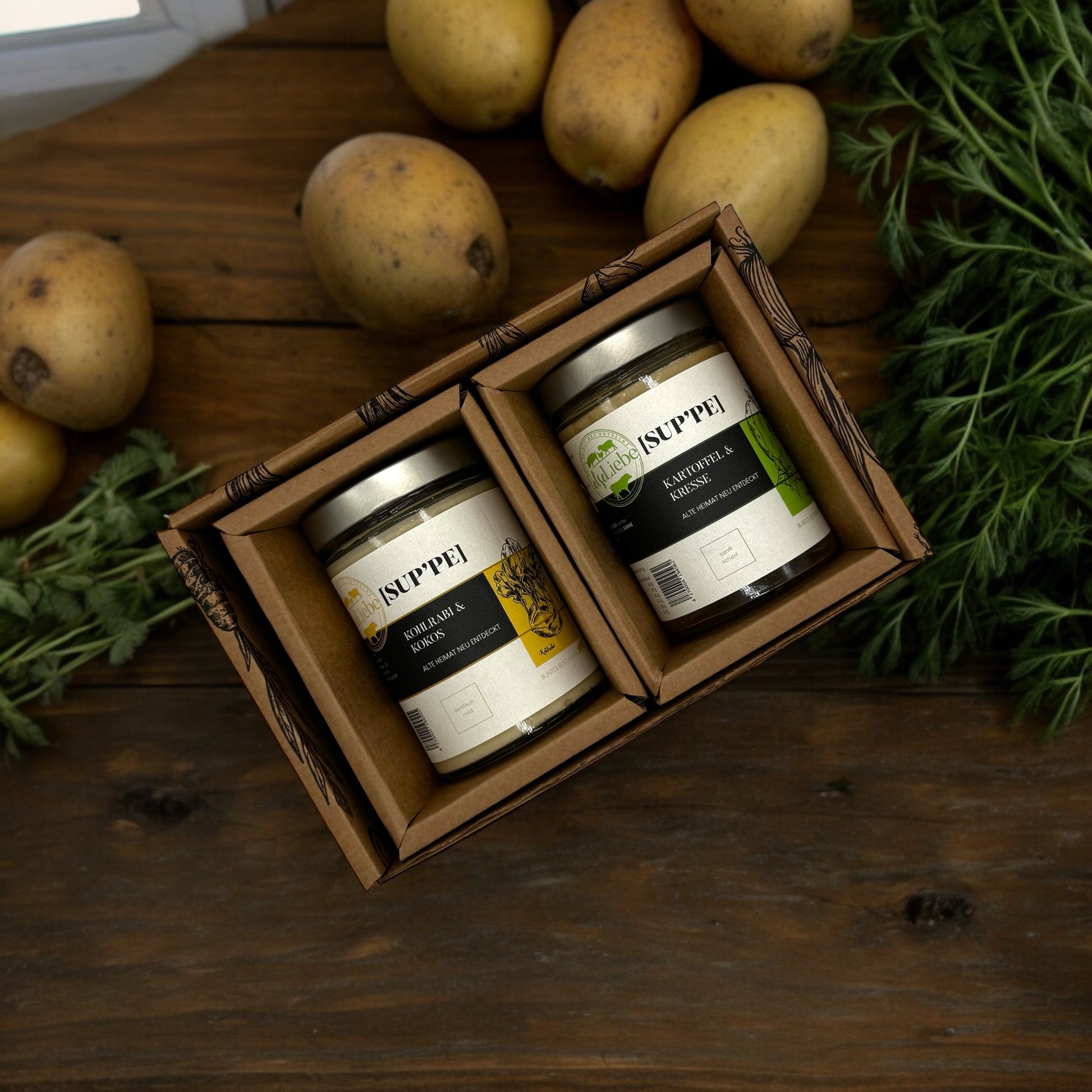 Auf dem Bild liegt eine kleine Genussbox der Marke elseLiebe auf einer Arbeitsplatte. Im Hintergrund sind Kräuter und Kartoffeln zu sehen. Die Sorten sind Kartoffel & Kresse und Kohlrabi & Kokos.