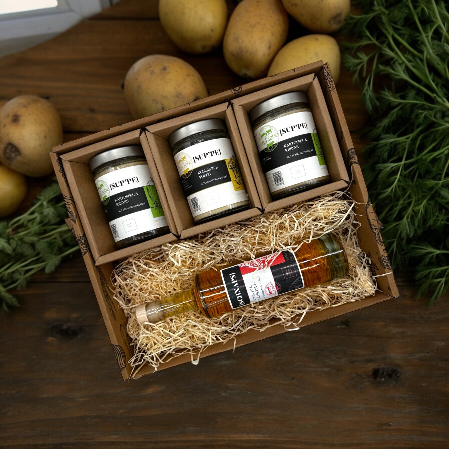 Eine Genussbox auf einem Tisch. Im Hintergrund sind Kartoffeln und Kräuter zu sehen. In der Box befinden sich drei Gläser mit Suppe. Die Sorten Kartoffel & Kresse und Kohlrabi & Kokos sind zu erkennen. Diese Suppen sind von der Marke elseLiebe und vom Bunten Bentheimer Schwein gemacht. Darunter liegt auf etwas Holzwolle eine Flasche Schnaps von der Marke elseLiebe. Es ist ein Aquavitlikör mit Grapefruit.