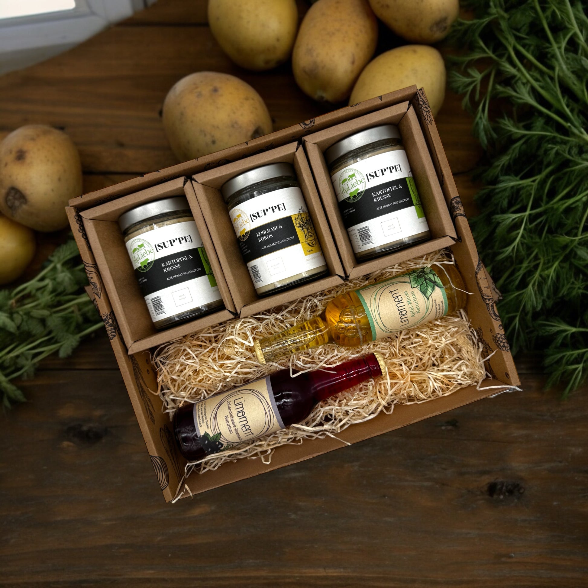 Eine Genussbox auf einem Tisch. Im Hintergrund sind Kartoffeln und Kräuter zu sehen. In der Box befinden sich drei Gläser mit Suppe. Die Sorten Kartoffel & Kresse und Kohlrabi & Kokos sind zu erkennen. Diese Suppen sind von der Marke elseLiebe und vom Bunten Bentheimer Schwein gemacht. Darunter liegen auf etwas Holzwolle zwei Flaschen Limoment Limo. Es sind die Sorten Apfel-Minze und Johannisbeere-Rosmarin.