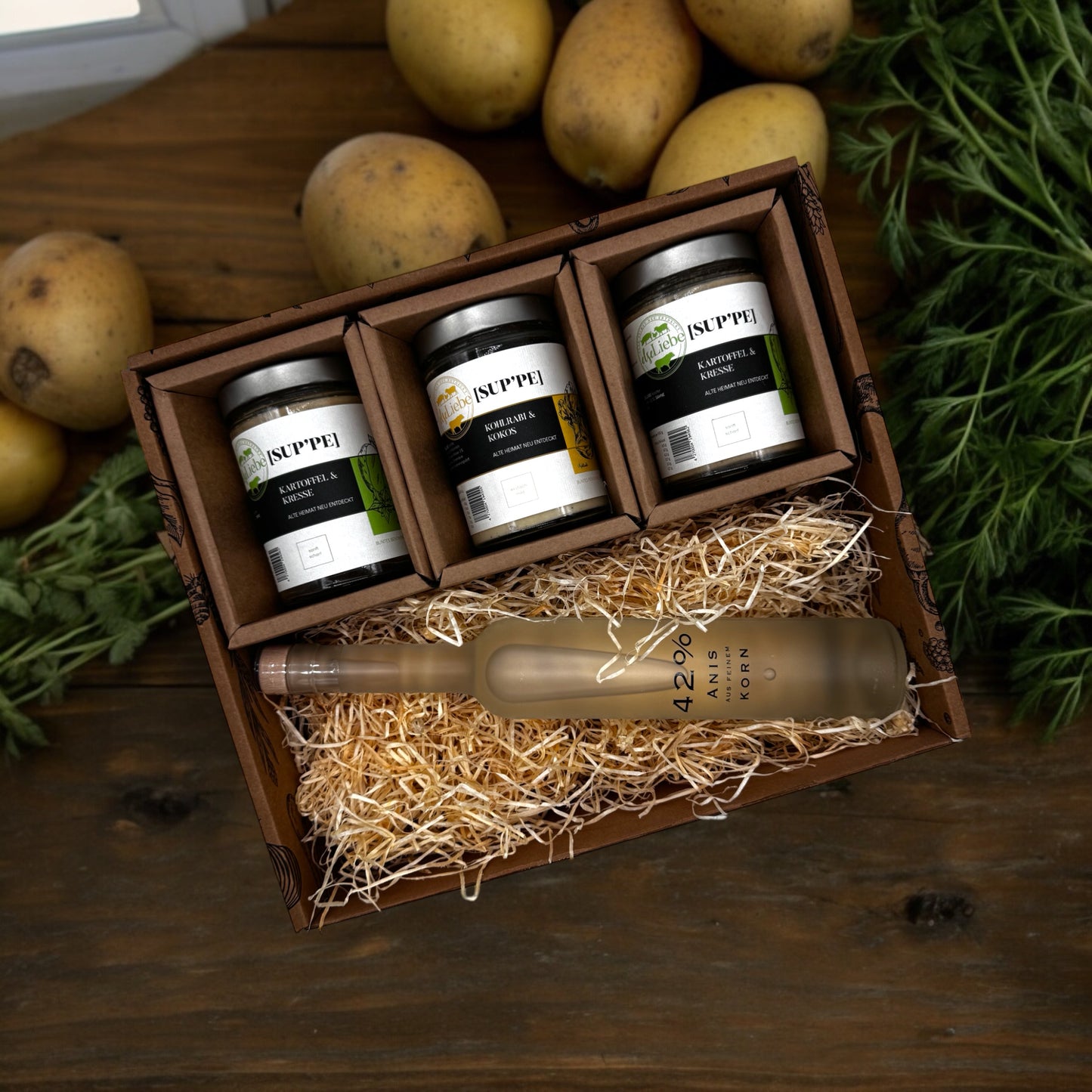 Eine Genussbox auf einem Tisch. Im Hintergrund sind Kartoffeln und Kräuter zu sehen. In der Box befinden sich drei Gläser mit Suppe. Die Sorten Kartoffel & Kresse und Kohlrabi & Kokos sind zu erkennen. Diese Suppen sind von der Marke elseLiebe und vom Bunten Bentheimer Schwein gemacht. Darunter liegt auf etwas Holzwolle eine Flasche Schnaps eines Anislikörs mit Korn.