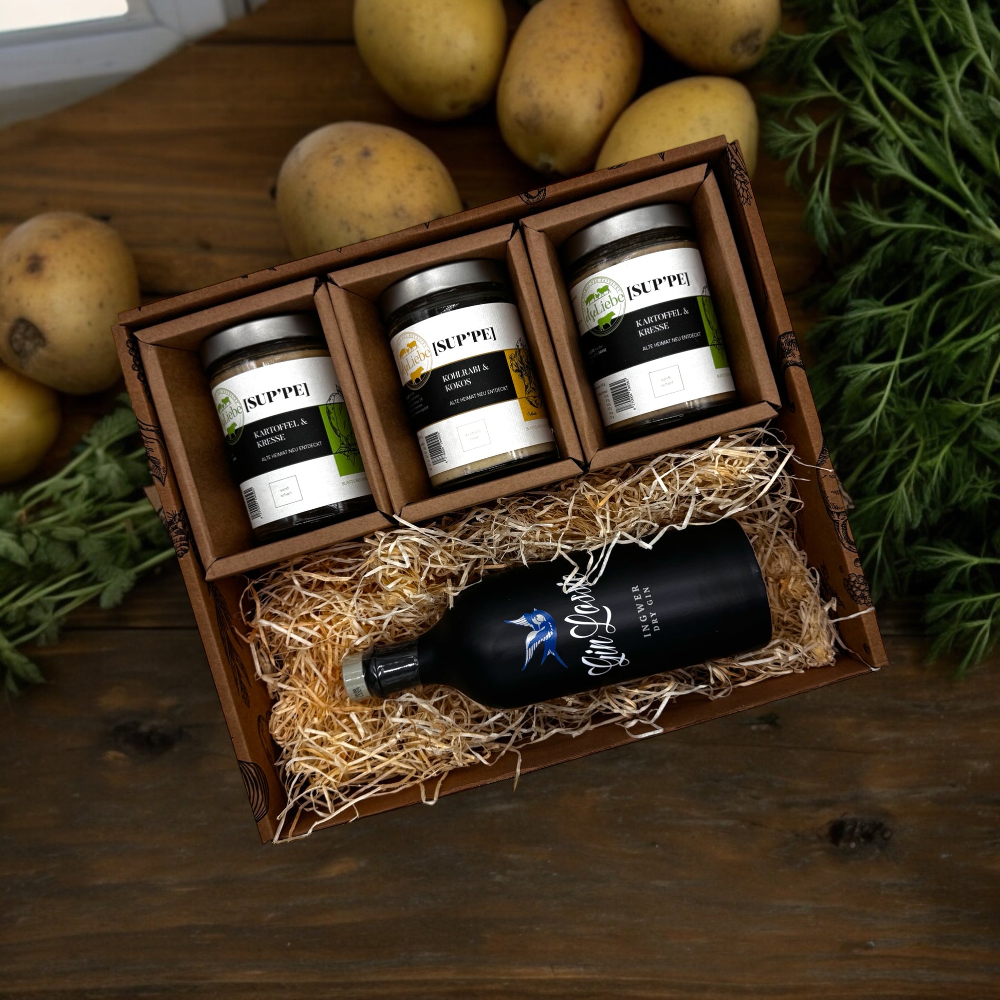 Eine Genussbox auf einem Tisch. Im Hintergrund sind Kartoffeln und Kräuter zu sehen. In der Box befinden sich drei Gläser mit Suppe. Die Sorten Kartoffel & Kresse und Kohlrabi & Kokos sind zu erkennen. Diese Suppen sind von der Marke elseLiebe und vom Bunten Bentheimer Schwein gemacht. Darunter liegt auf etwas Holzwolle eine Flasche vom Gin Lossie der Sorte Dry Gin Ingwer.