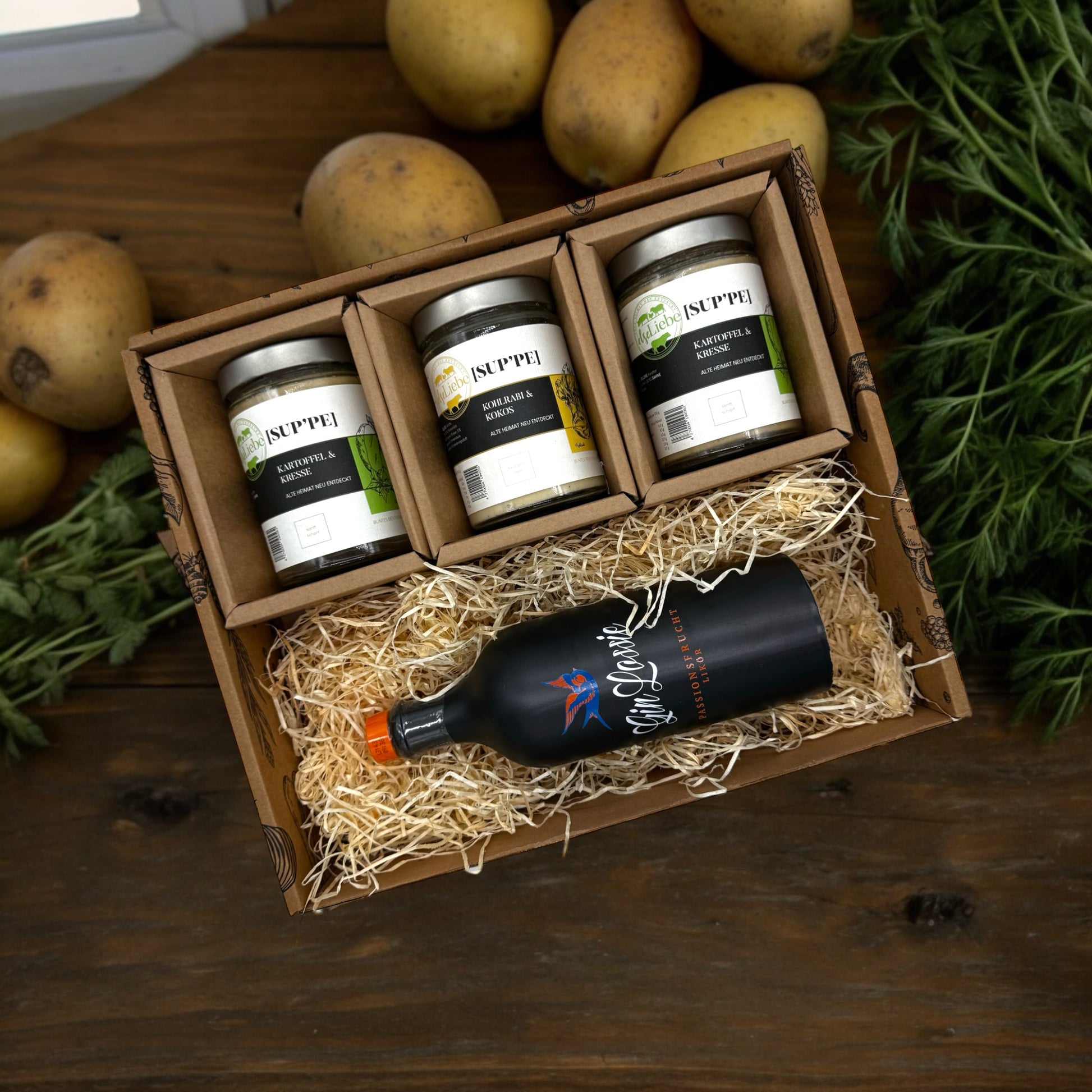 Eine Genussbox auf einem Tisch. Im Hintergrund sind Kartoffeln und Kräuter zu sehen. In der Box befinden sich drei Gläser mit Suppe. Die Sorten Kartoffel & Kresse und Kohlrabi & Kokos sind zu erkennen. Diese Suppen sind von der Marke elseLiebe und vom Bunten Bentheimer Schwein gemacht. Darunter liegt auf etwas Holzwolle eine Flasche vom Gin Lossie der Sorte Passionsfrucht.